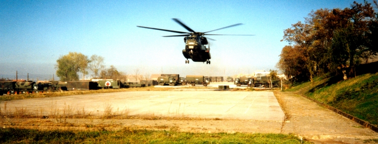 Ein planierter Platz an einem Erdhang; Fahrzeuge mit Rotem Kreuz; in der Mitte landet ein Helikopter