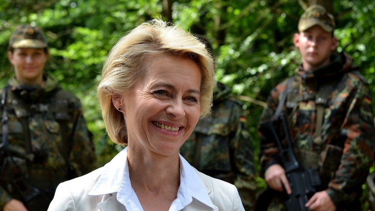 Verteidigungsministerin Ursula von der Leyen lächelt in die Kamera. Im Hintergrund links eine lächelnde deutsche Soldatin, rechts ein Soldat mit Waffe und gelangweiltem Blick